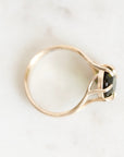 A gold ring with a bezel-set black gemstone, photographed from an angled top-down view on a light marble surface.