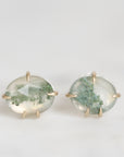 A pair of oval stud earrings featuring milky white stones with green inclusions, held in gold prongs against a light gray background.