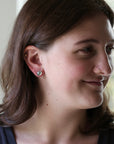 A close-up of a woman with brown hair wearing a small silver hoop earring with a green heart-shaped crystal on her earlobe, along with a gold nose ring.