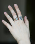 A close-up of a hand wearing a gold ring with a clear round blue with a small blue geometric tattoo on the middle finger against a dark blurred background.