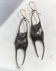 A pair of sculptural dangle earrings with dark oxidized metal bodies and gold-tone hook ear wires, resting on a light marble surface.