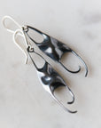 A pair of sculptural dangle earrings with organic, flowing shapes in bright silver and dark oxidized black, resting on a light marble surface.