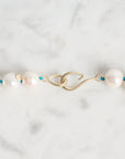A close-up of a pearl necklace with white round pearls threaded on a gold-toned chain, featuring a gold clasp and small blue accents, laid on a light marble surface.