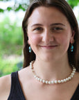 A woman with long brown hair smiling, wearing a white pearl necklace and matching drop earrings with blue stones, set against a soft green outdoor background.