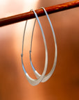 Large Facet Hoops in silver hanging from a wood stick, set against an orange background- Hannah Blount Jewelry