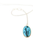 A turquoise pendant with a scrimshaw mermaid illustration on a long silver chain.