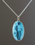 A turquoise pendant with a scrimshaw mermaid illustration on a long silver chain.