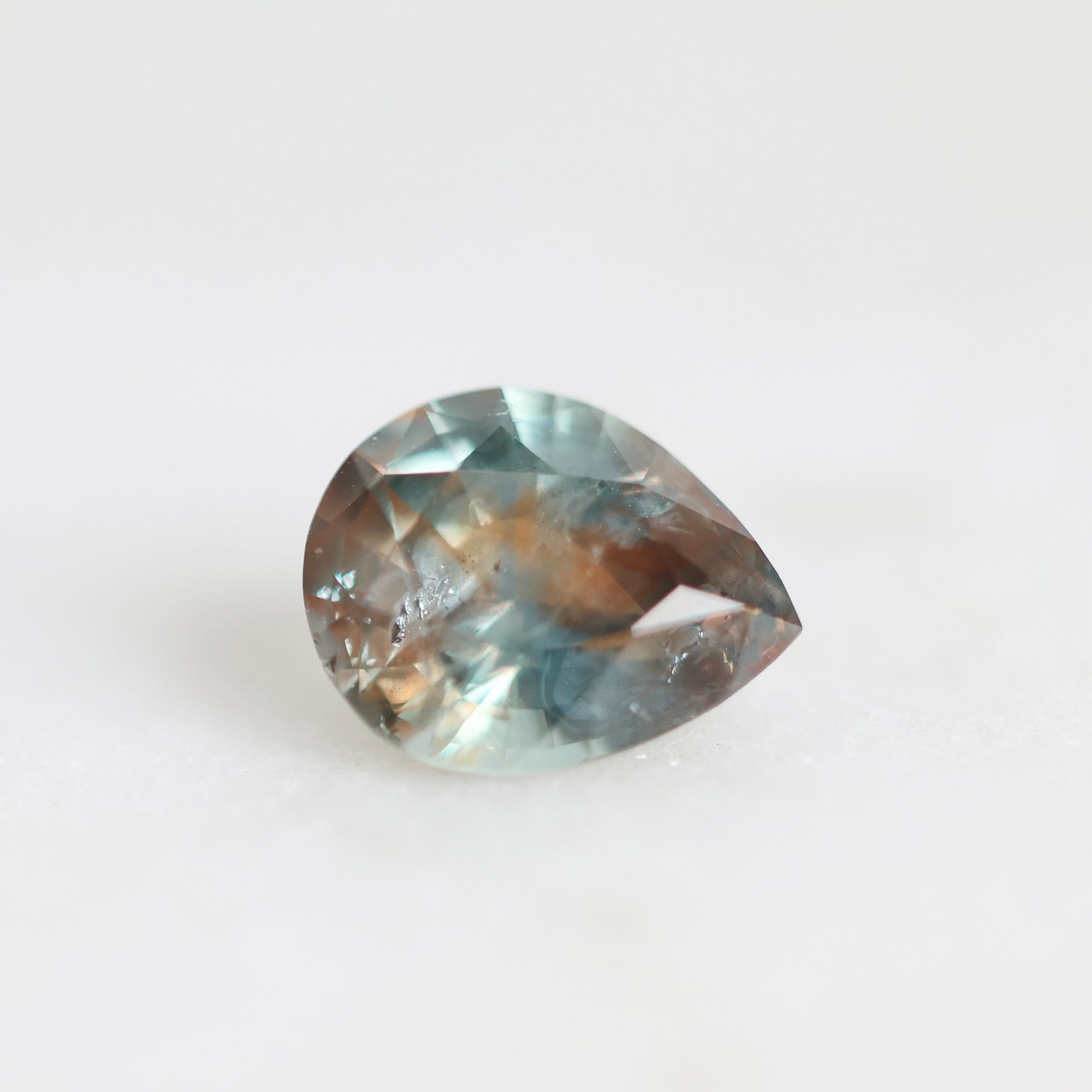 A teardrop-shaped faceted gemstone with a gradient of blue, teal, and brown tones, resting on a plain white surface.