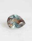 A teardrop-shaped faceted gemstone with a gradient of blue, teal, and brown tones, resting on a plain white surface.