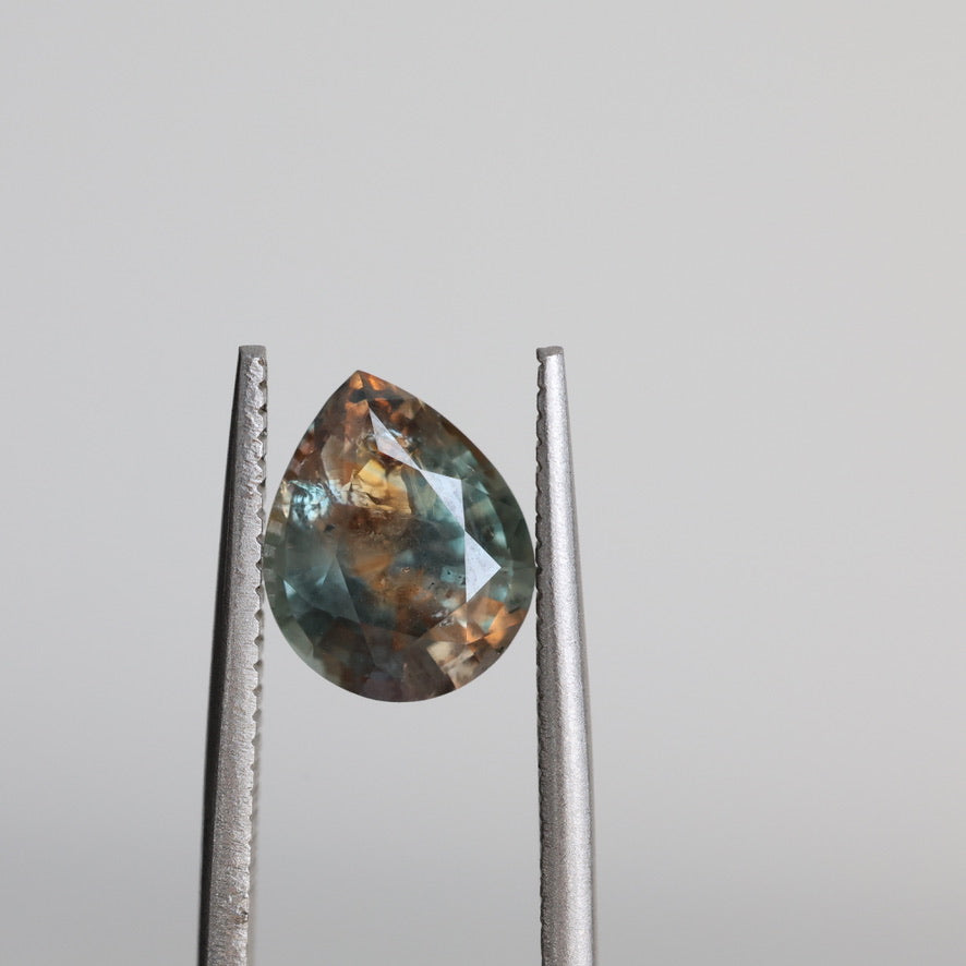 A teardrop-shaped faceted gemstone held between metal tweezers against a plain light gray background.