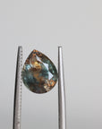 A teardrop-shaped faceted gemstone held between metal tweezers against a plain light gray background.