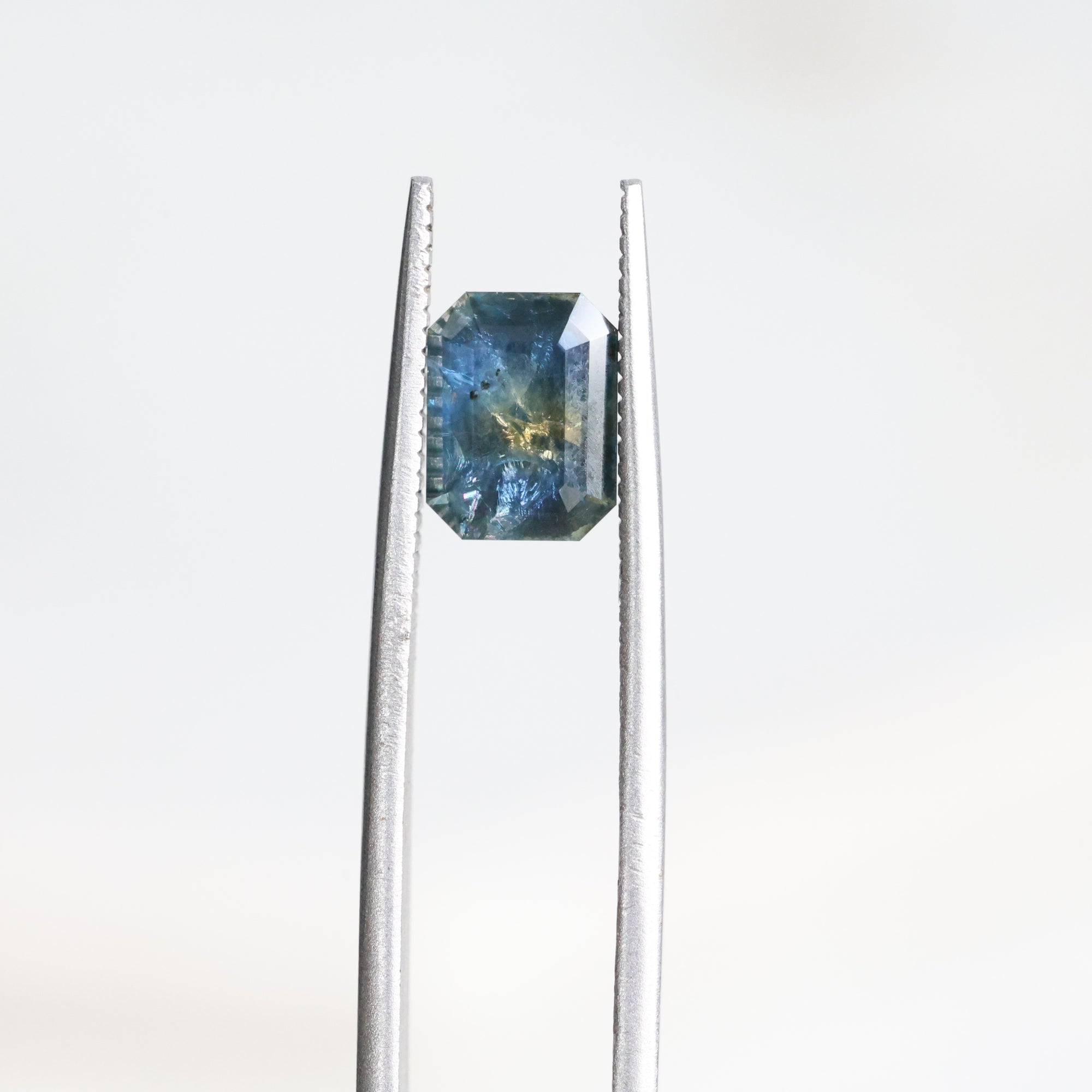 A loose rectangular blue gemstone with internal inclusions, held between silver metal tweezers against a plain white background.