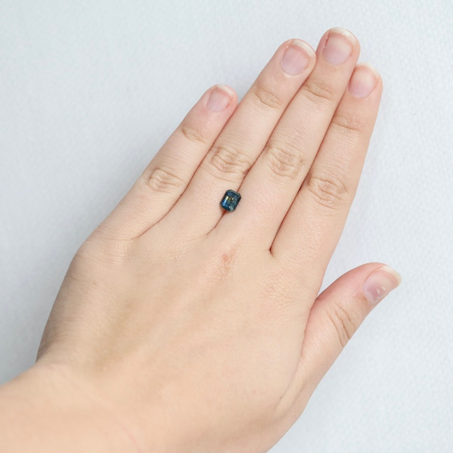 A close-up of a hand wearing a small rectangular dark blue gemstone ring on the ring finger, set against a plain light gray background.