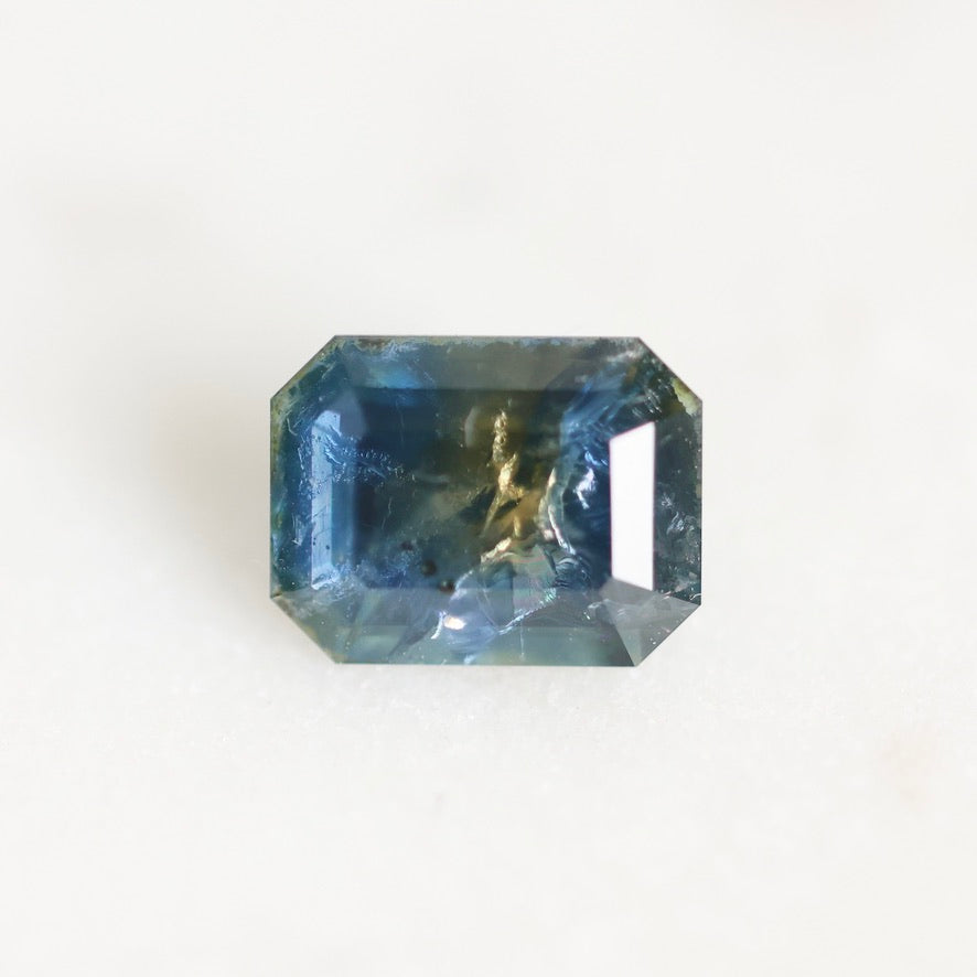 A rectangular emerald-cut blue gemstone with natural inclusions and a gold-colored matrix core, photographed on a clean white background.