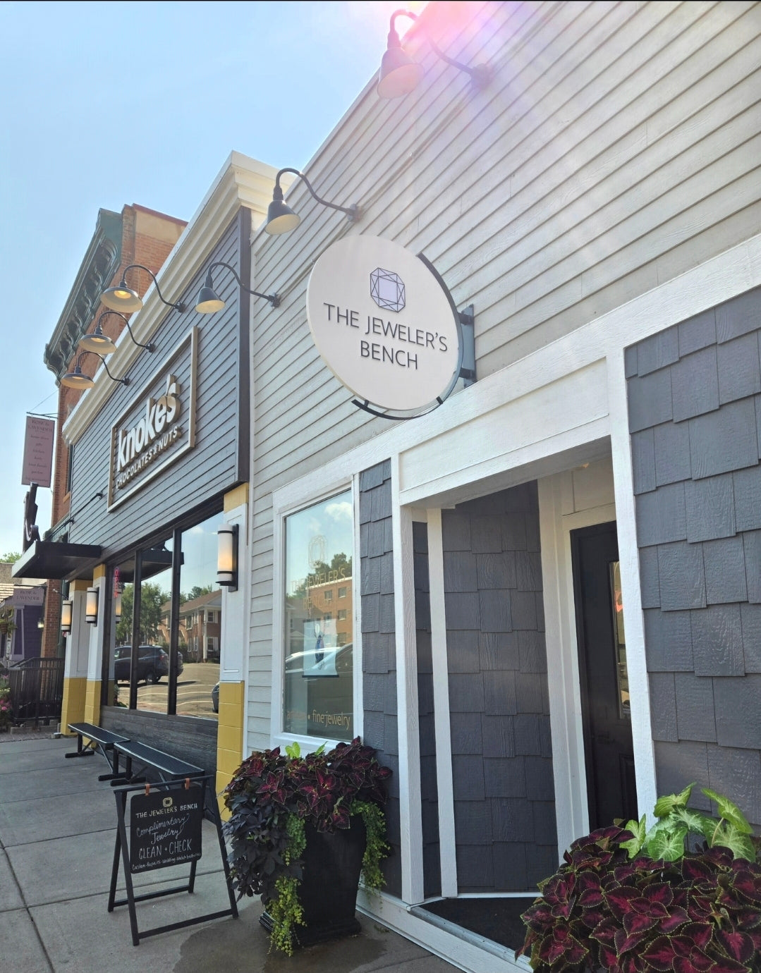 The exterior of 'The Jeweler's Bench' storefront, featuring a white siding facade with dark gray shingled panels, a round white sign, and potted plants on the sidewalk.