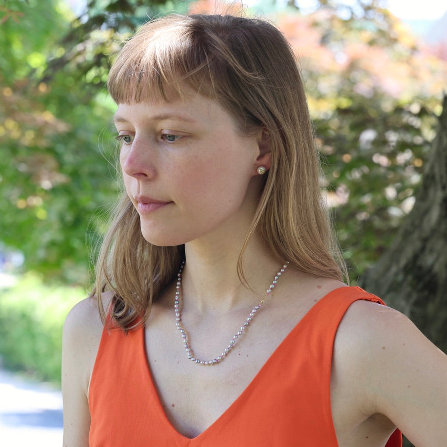 Woman wearing an orange top and pearl necklace outdoors with greenery in the background