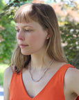 Woman wearing an orange top and pearl necklace outdoors with greenery in the background