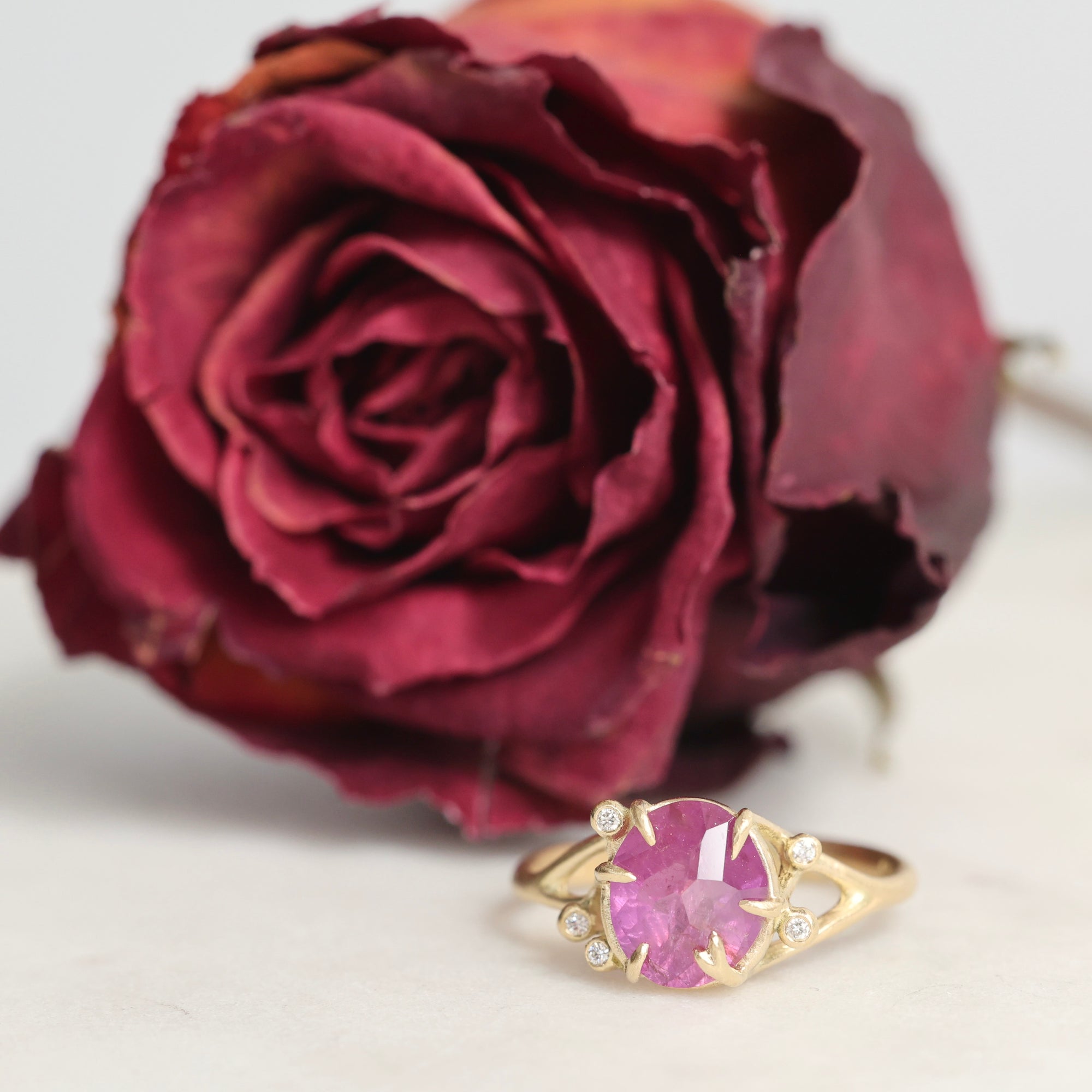 Gold ring with a pink gemstone next to a red rose on a light background