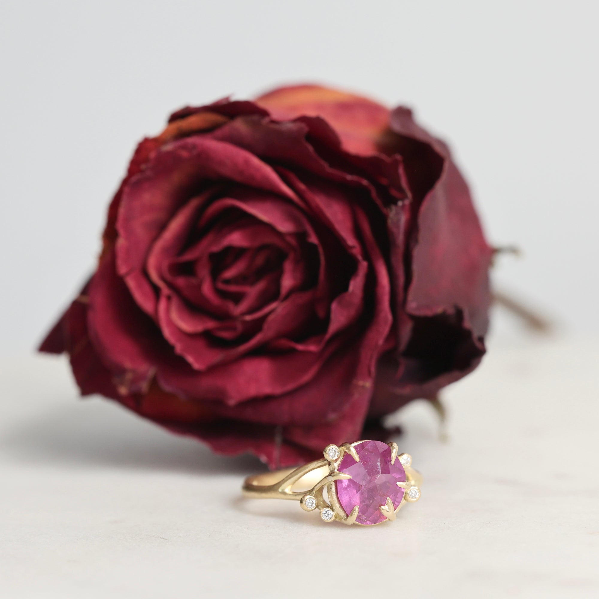 Gold ring with a pink gemstone next to a dried red rose on a light background
