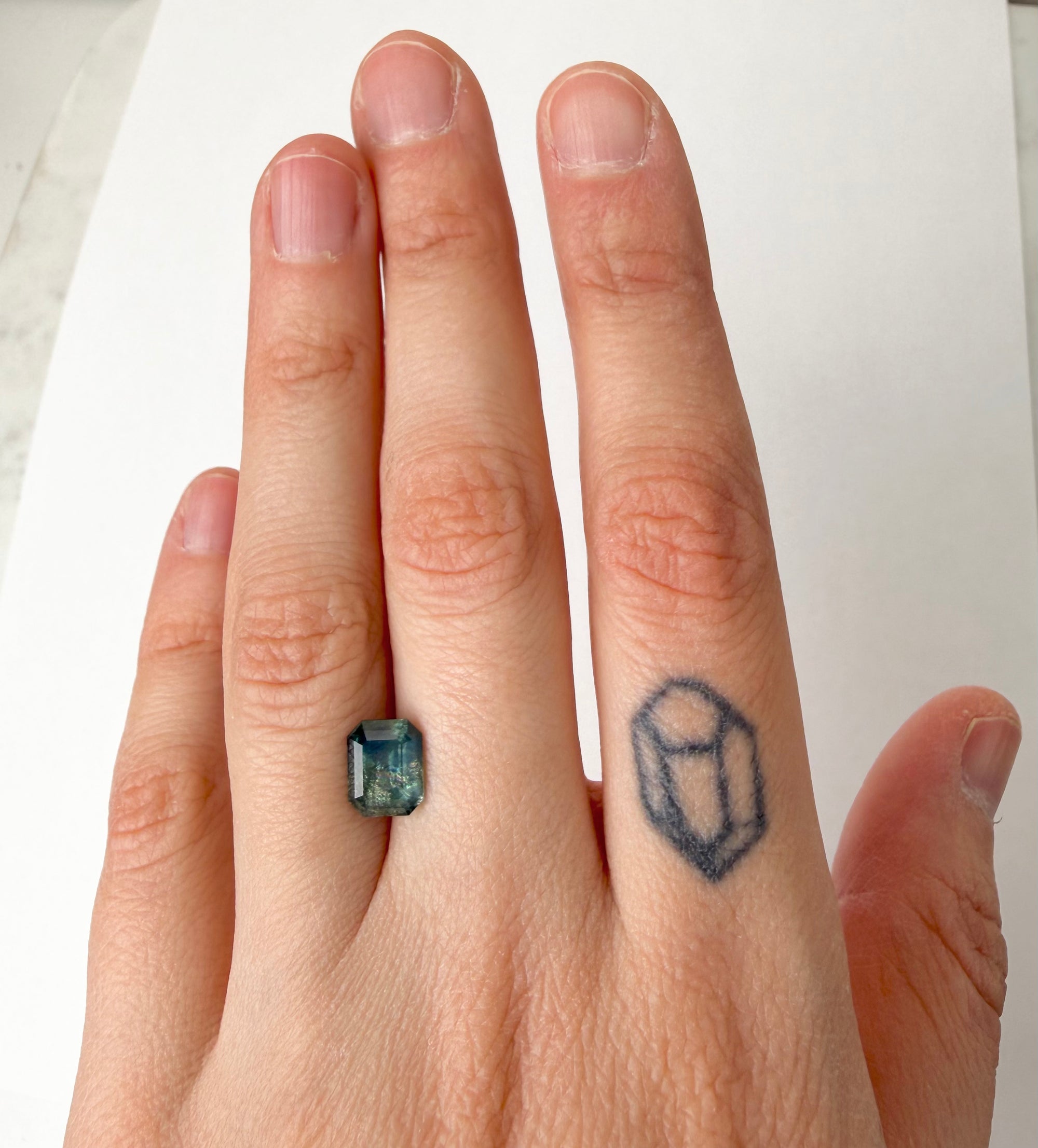 Hand with a ring featuring an emerald-cut stone and a small tattoo on the ring finger.