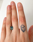 Hand with a ring featuring an emerald-cut stone and a small tattoo on the ring finger.