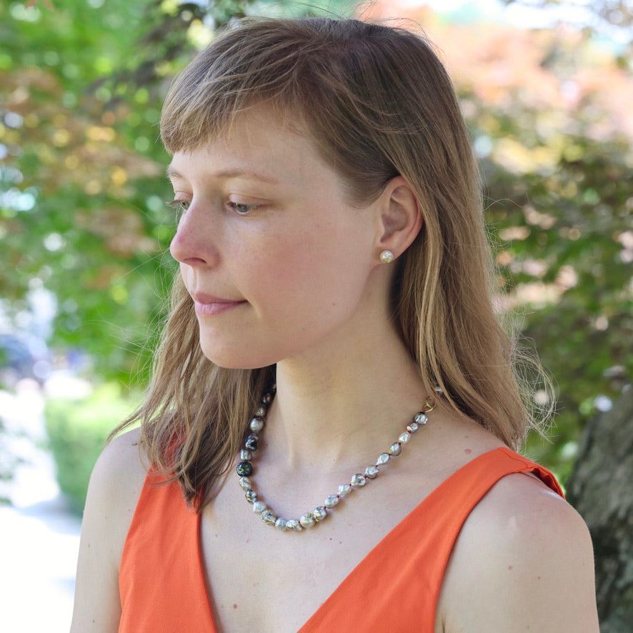 Woman wearing an orange top and pearl necklace outdoors with blurred greenery in the background