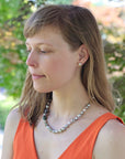 Woman wearing an orange top and pearl necklace outdoors with blurred greenery in the background