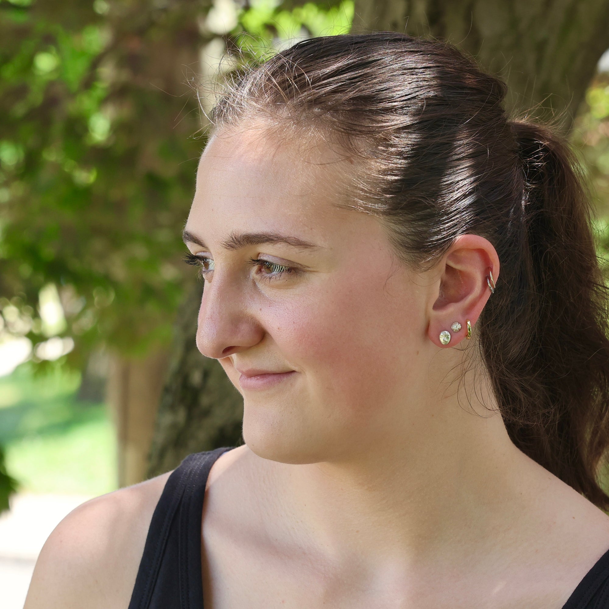 Woman with a ponytail wearing earrings, standing outdoors with trees in the background