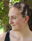 Woman with a ponytail wearing earrings, standing outdoors with trees in the background