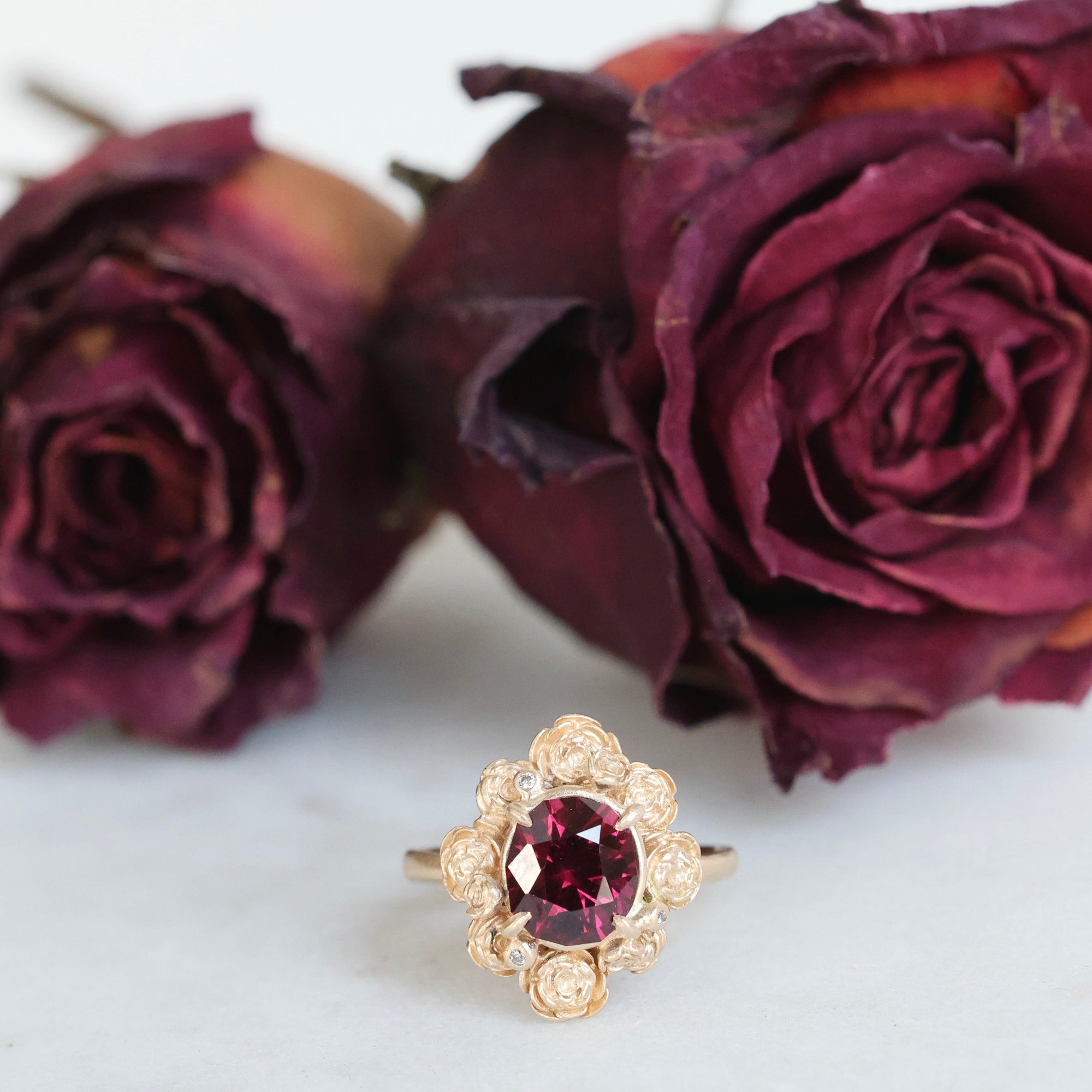 Gold ring with a red gemstone in front of dried purple roses on a white background