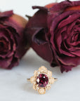Gold ring with a red gemstone in front of dried purple roses on a white background