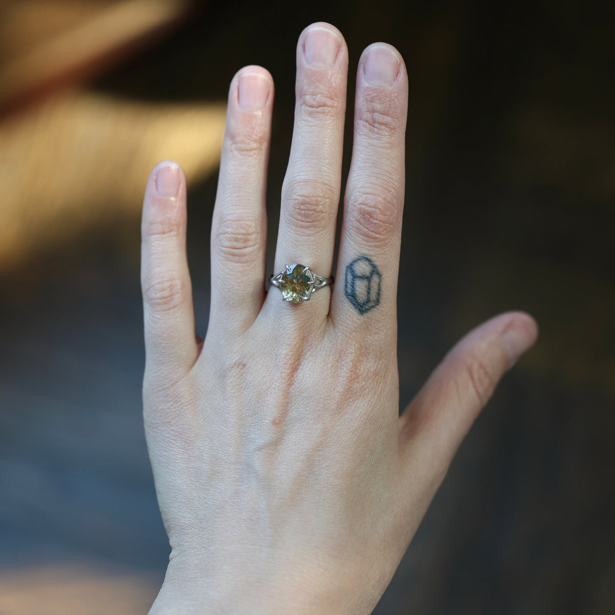 A close-up of a person's hand wearing a silver ring with a green gemstone, with a small geometric tattoo on one of the fingers.