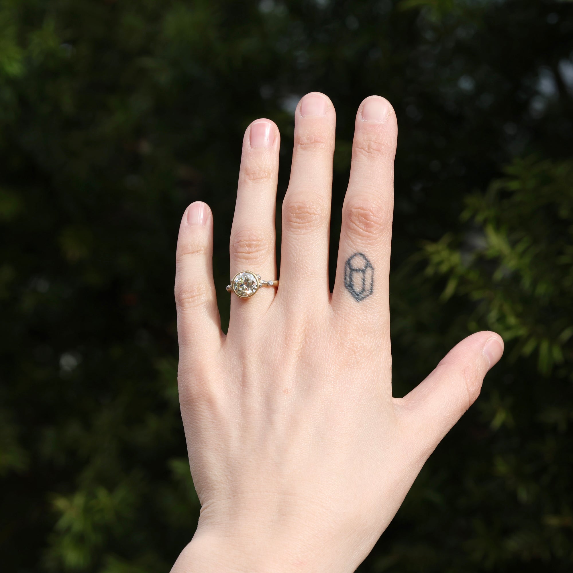 Hand with a diamond ring on a finger and a small tattoo, against a blurred green background