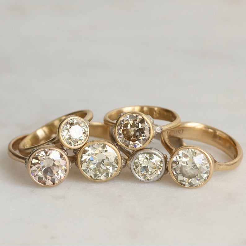 Five gold rings with round clear gemstones, arranged together on a light marble surface.