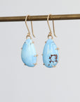 Pair of blue teardrop earrings on a gold earring stand against a light gray background