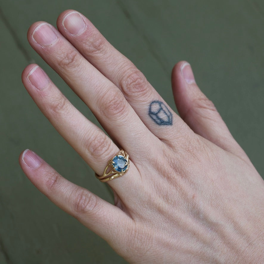 Hand wearing a gold ring with a blue gemstone on a neutral background