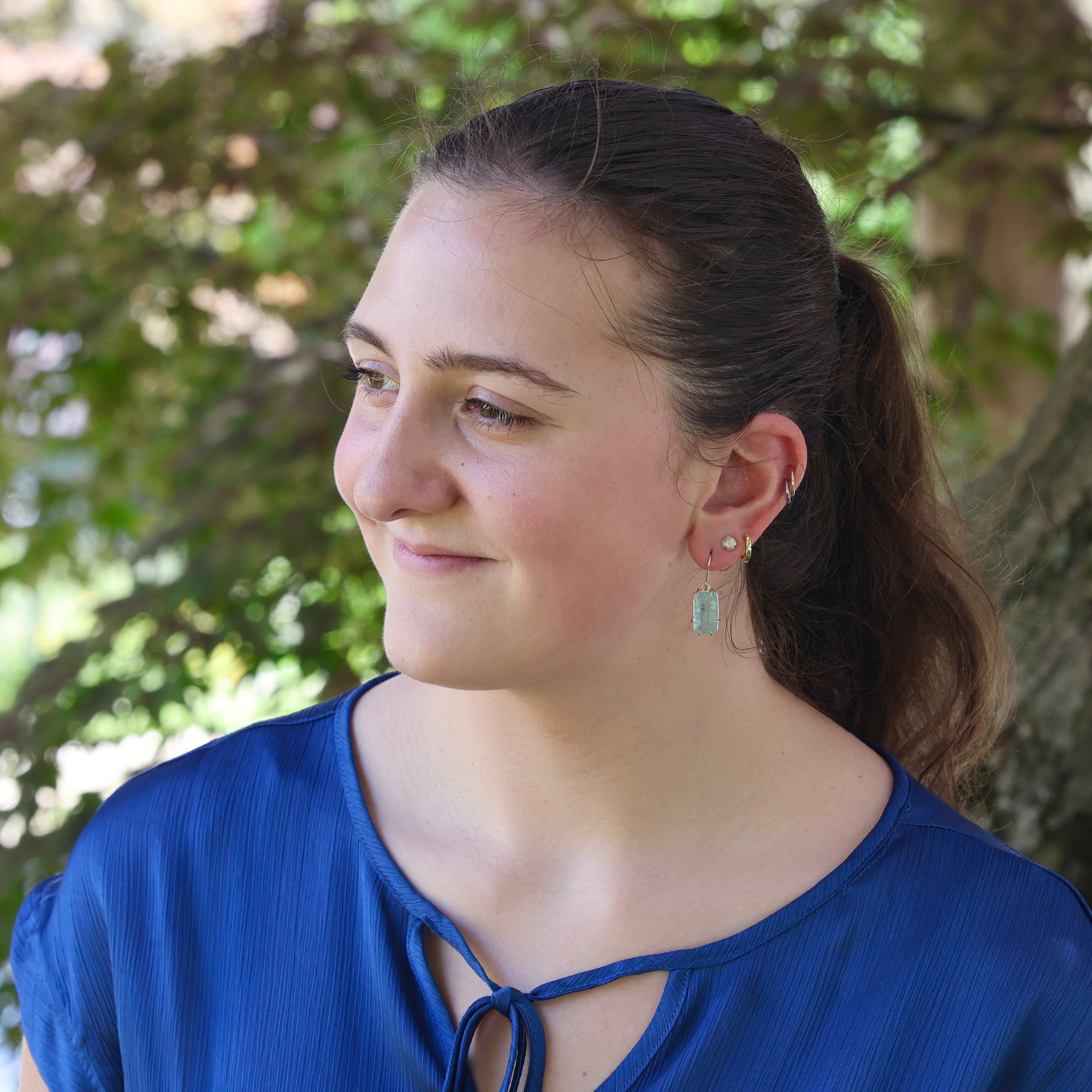 Woman wearing a blue top with earrings, standing outdoors with greenery in the background