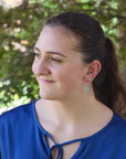Woman wearing a blue top with earrings, standing outdoors with greenery in the background