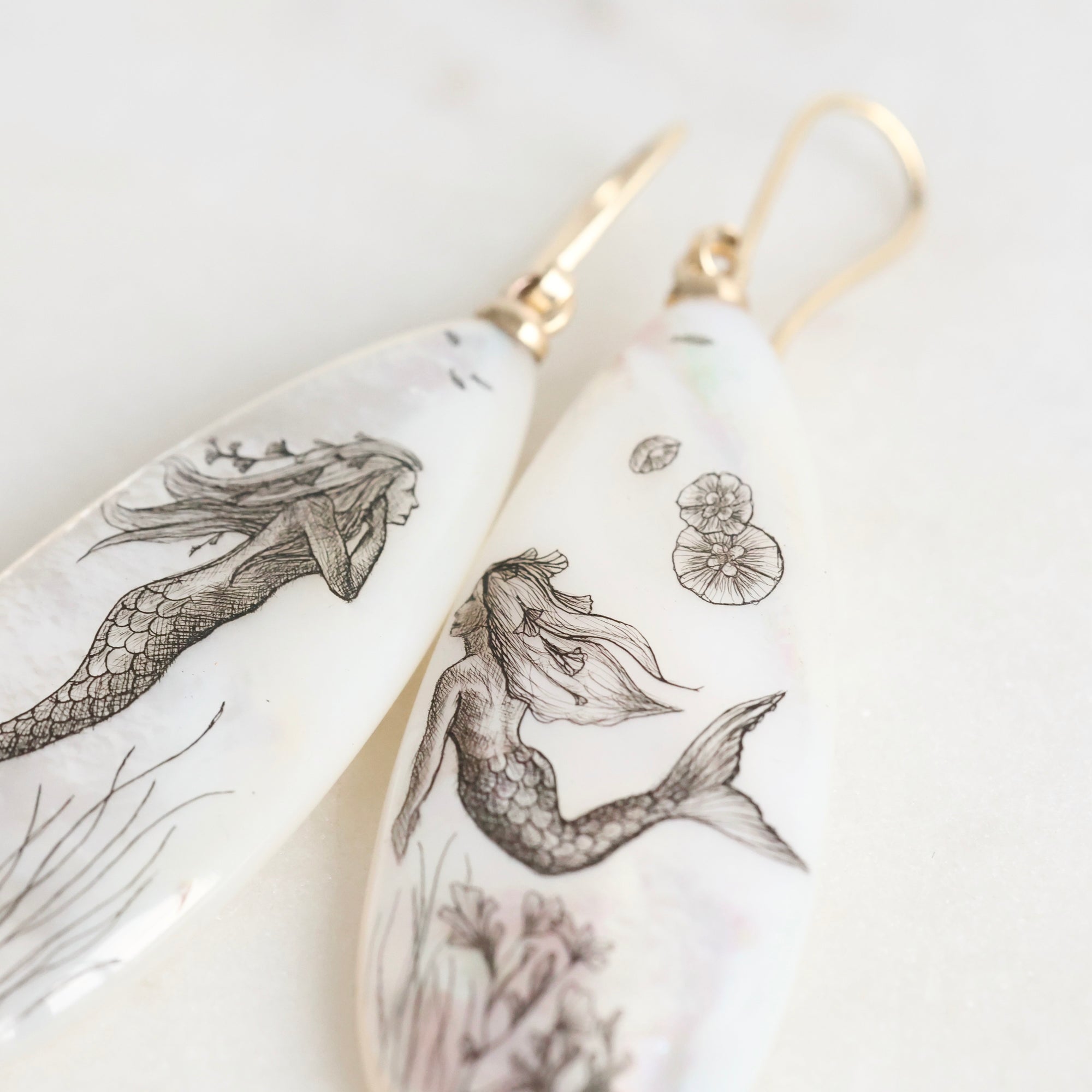 Teardrop-shaped earrings with mermaid illustrations on a light background