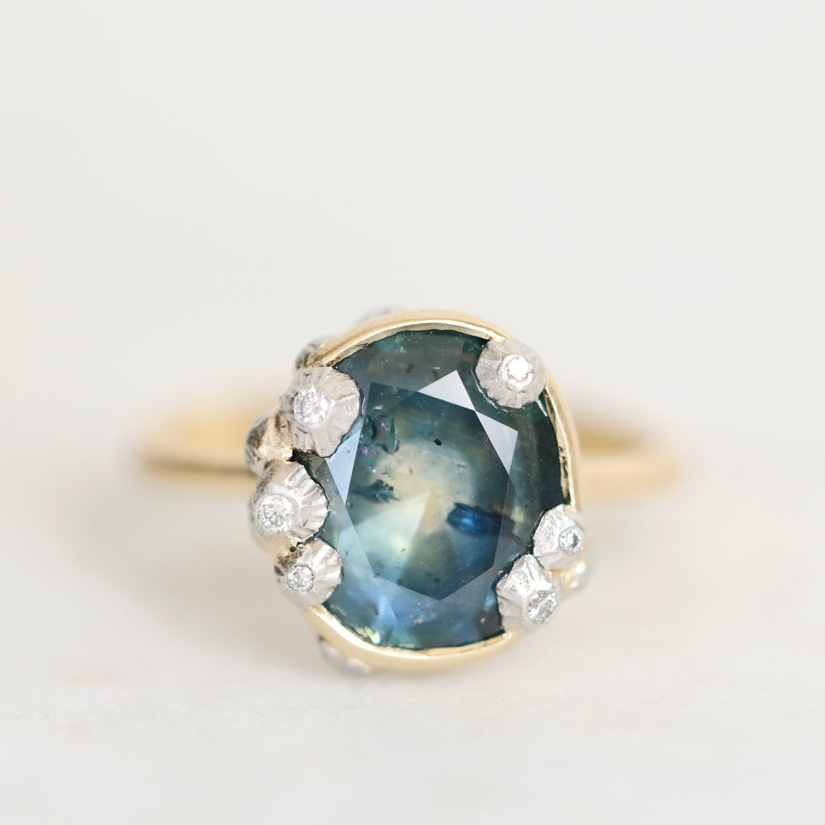 An antique-style ring featuring a large oval blue sapphire with inclusions, surrounded by small white diamonds in a silver setting, with a yellow gold band visible on either side, shot against a plain off-white background.
