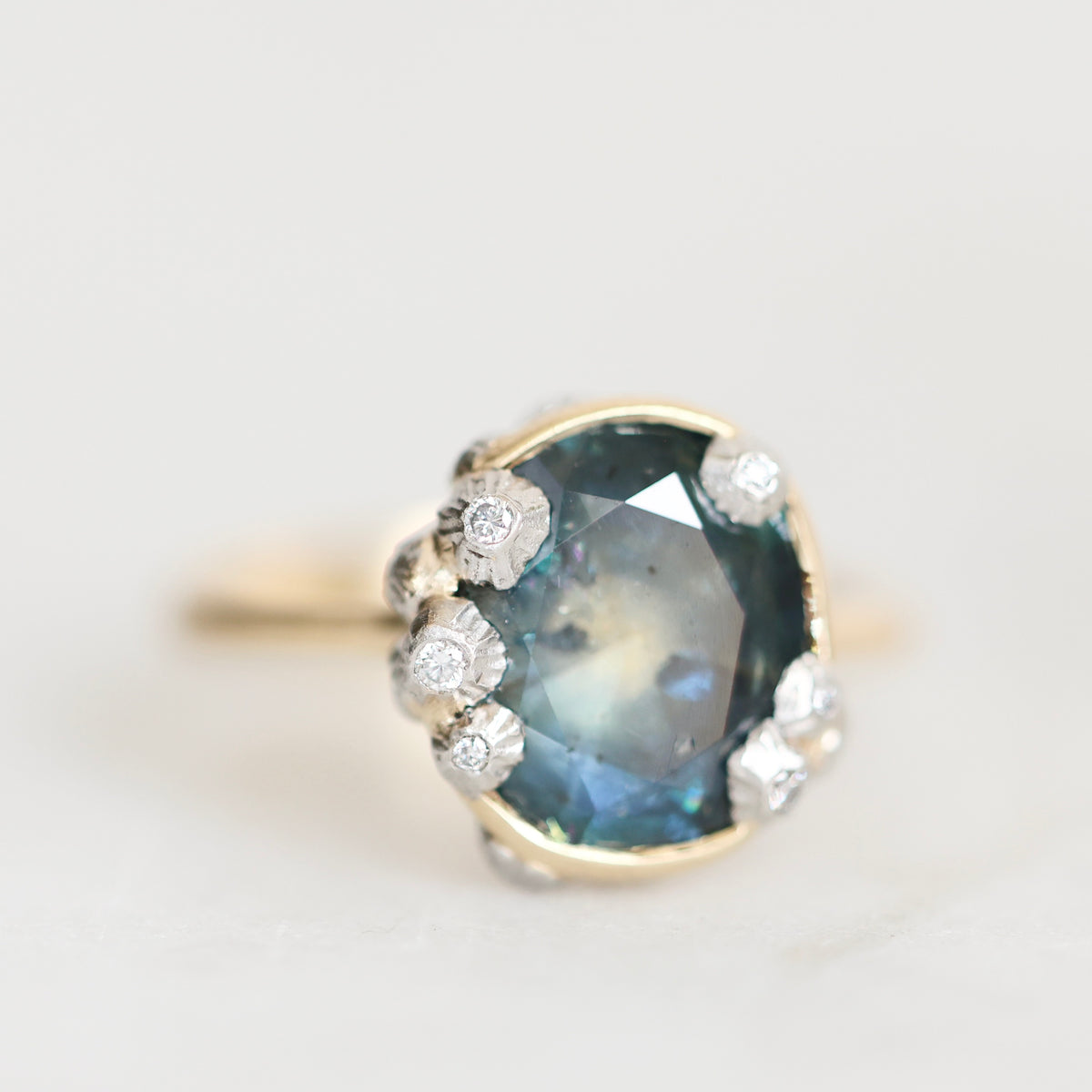 A vintage-style ring featuring a large oval blue gemstone in a gold-toned setting, accented with small white stones, photographed on a light neutral background.