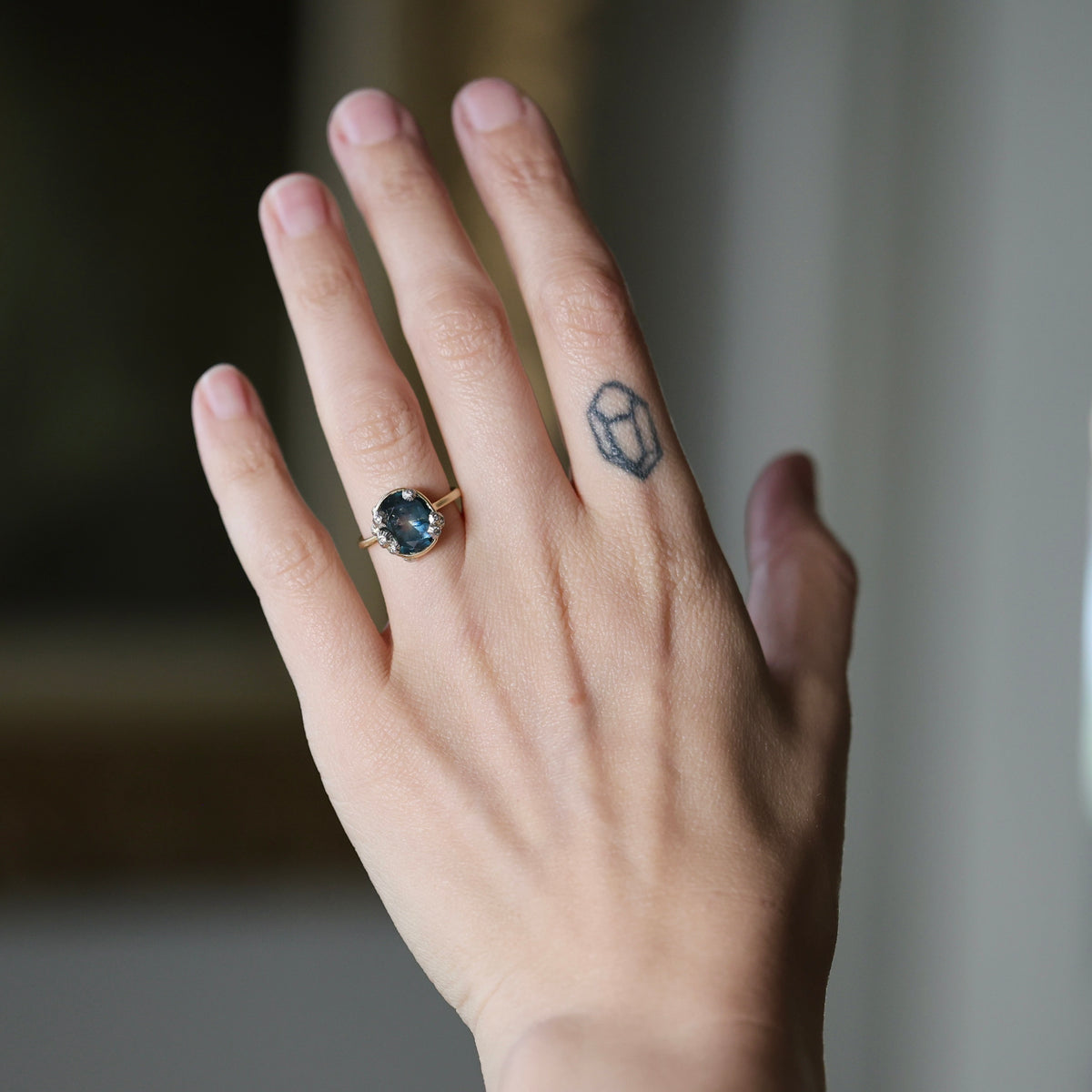 A close-up of a person's hand wearing a gold ring with a dark oval gemstone, alongside a small geometric tattoo on the side of the hand.