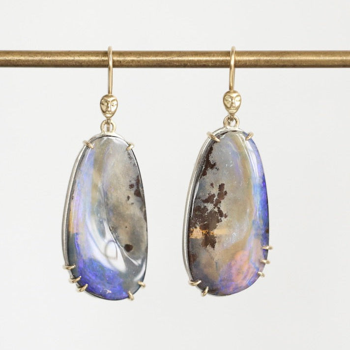 A pair of gold drop earrings with teardrop-shaped iridescent opal gemstones, hanging from a gold rod against a light gray background.