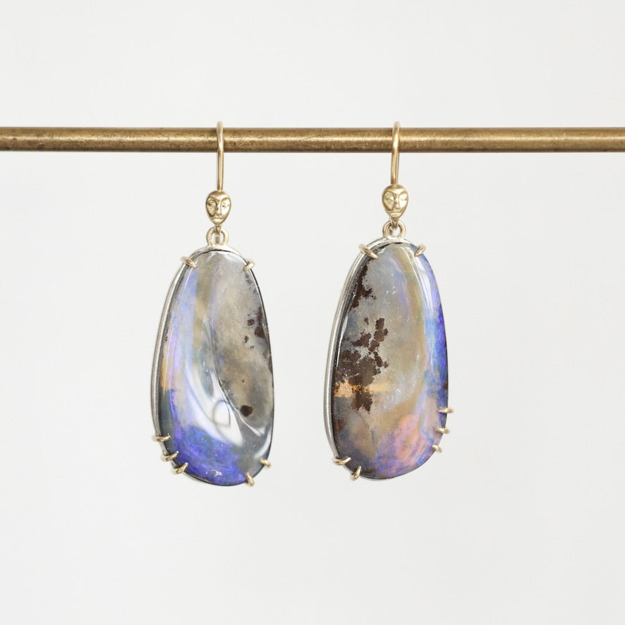 A pair of gold drop earrings with teardrop-shaped iridescent opal gemstones, hanging from a gold rod against a light gray background.
