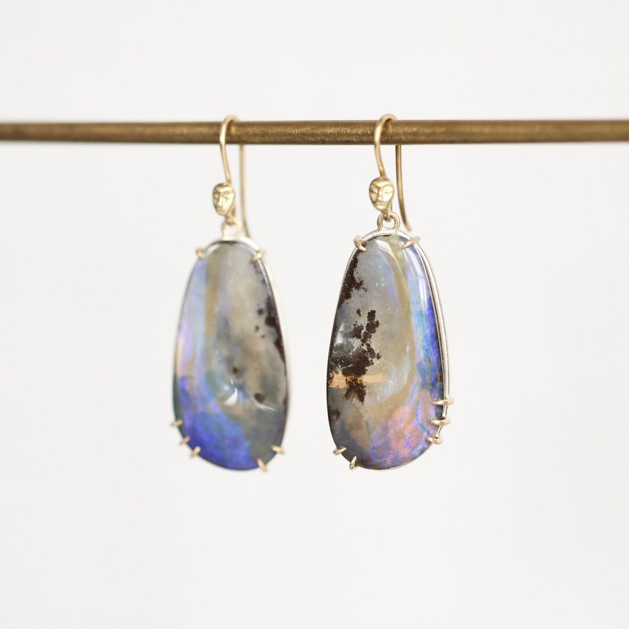 A pair of teardrop-shaped drop earrings with iridescent blue, purple, and brown opal-like stones in gold settings, hanging from a gold rod against a plain white background.