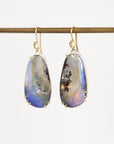 A pair of teardrop-shaped drop earrings with iridescent blue, purple, and brown opal-like stones in gold settings, hanging from a gold rod against a plain white background.
