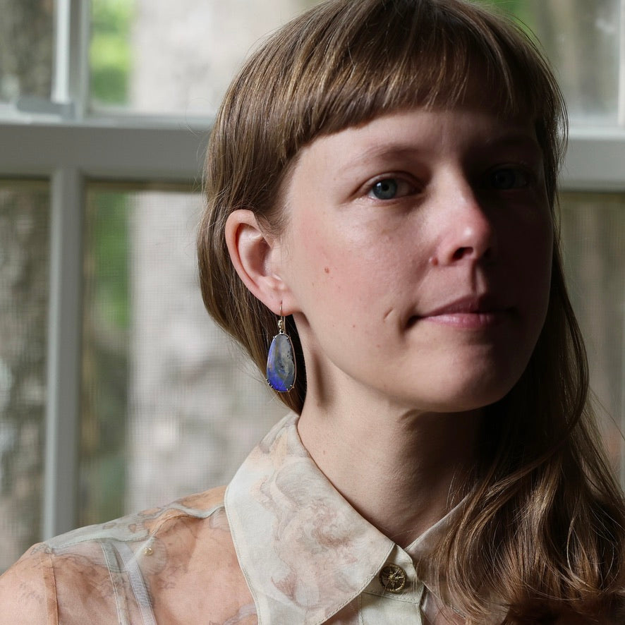 A close-up portrait of a woman with brown hair and bangs wearing a patterned cream blouse and a blue teardrop-shaped dangle earring with a gold-toned setting, posed in front of a soft-focus window.