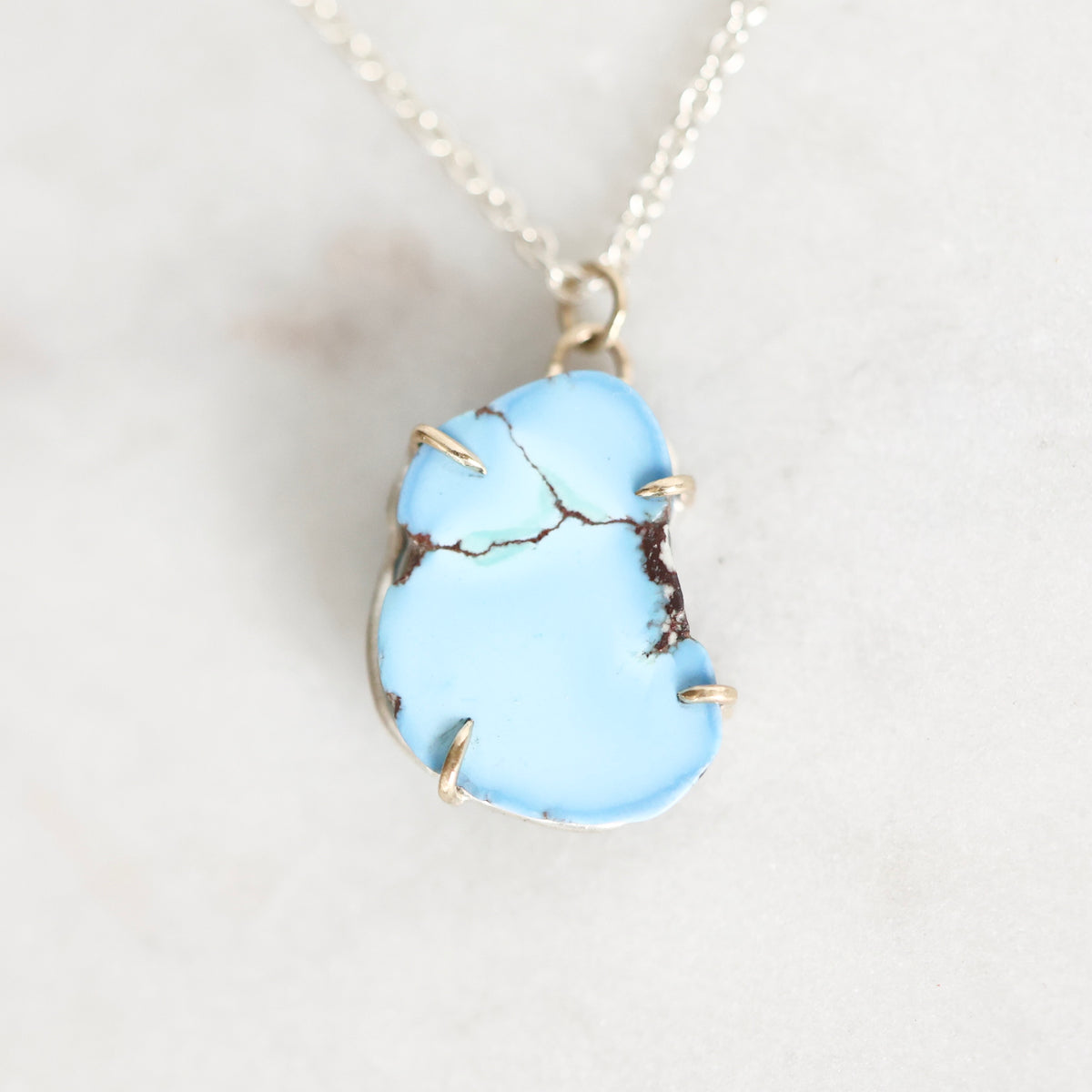 A light blue turquoise stone pendant necklace with gold wire wrapping and a silver chain, displayed on a light marble surface.