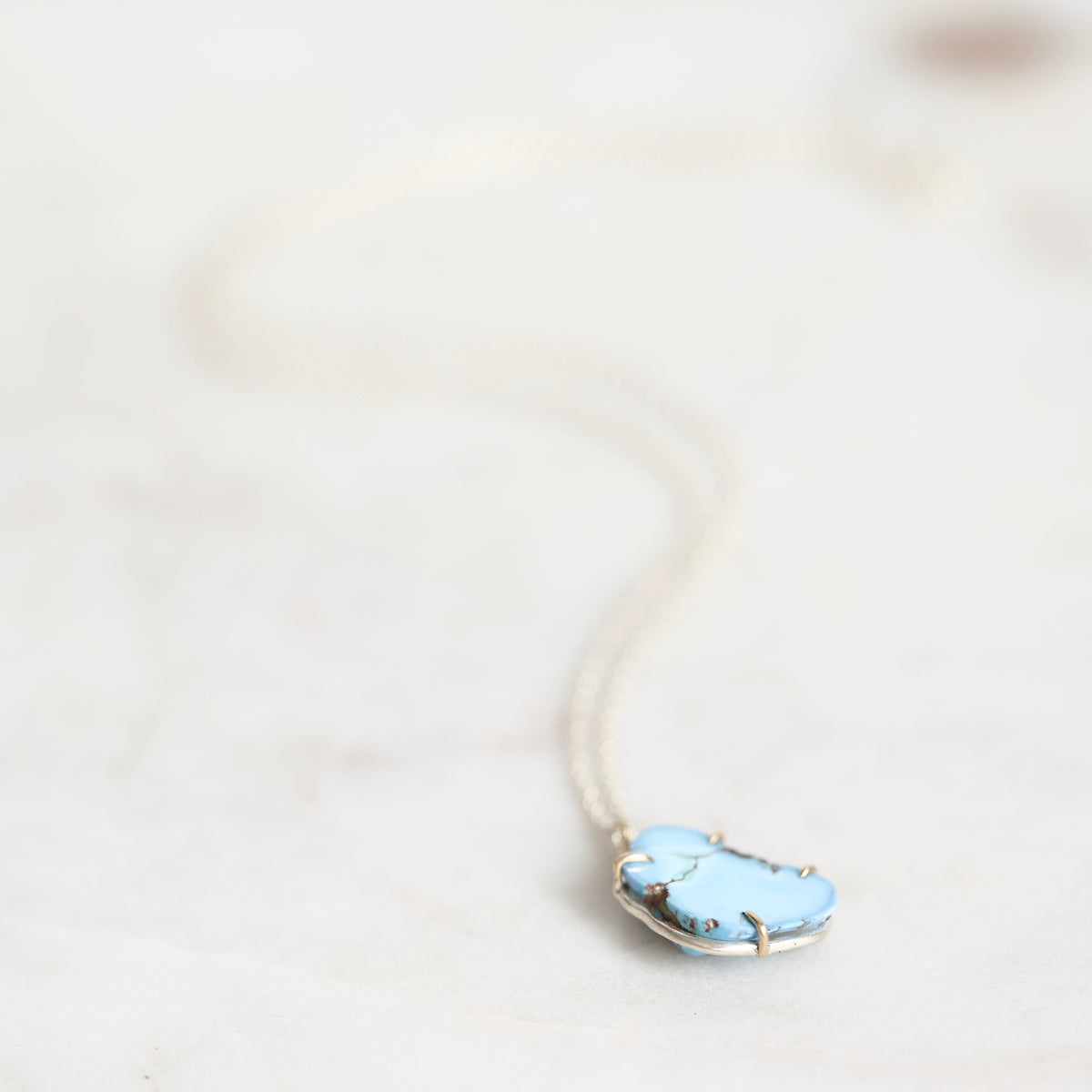 A delicate silver necklace with a light blue stone pendant, resting on a light marble surface.