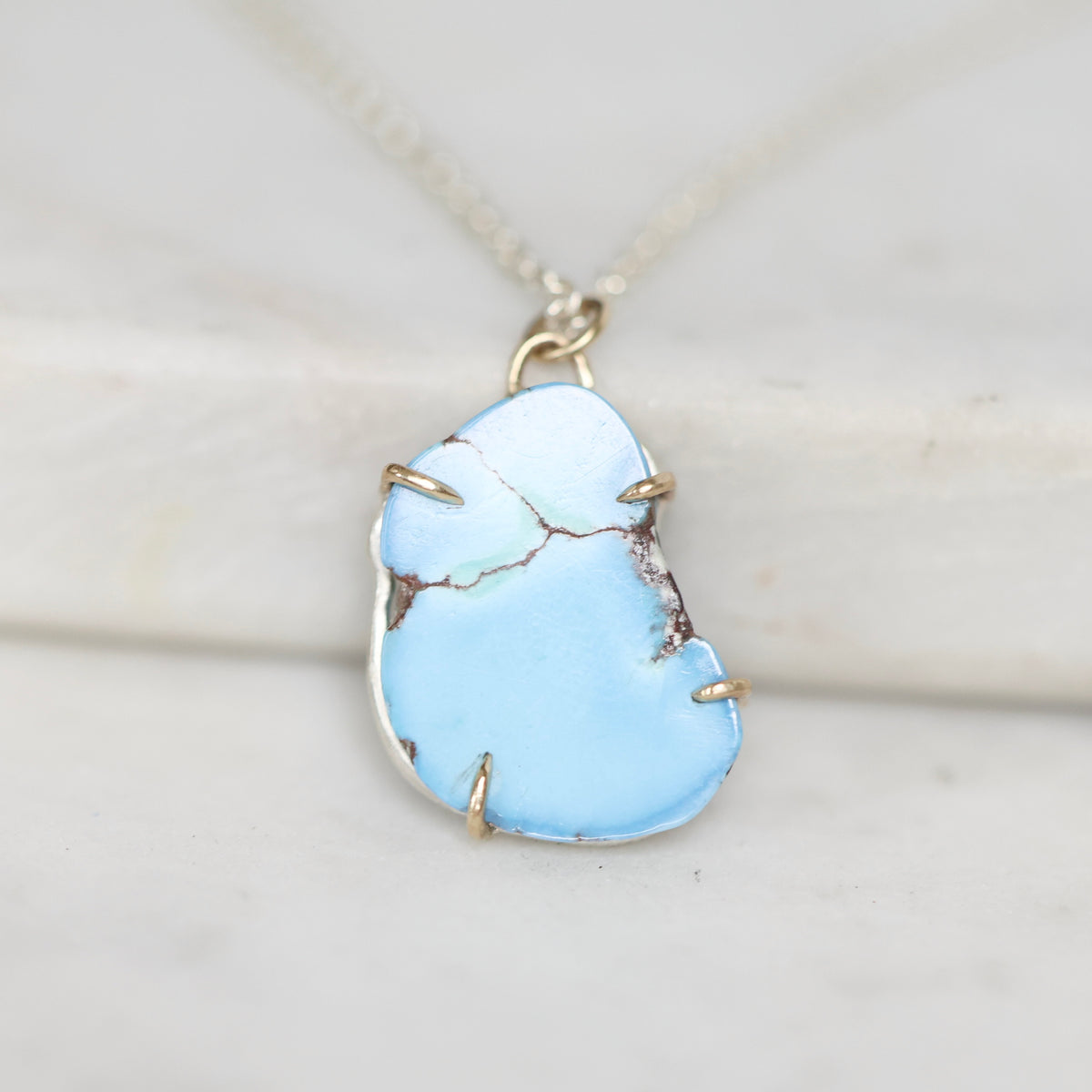 A close-up of a light blue turquoise stone pendant with natural matrix cracks, set in a gold-toned bezel and hanging from a silver-toned chain on a light marble surface.